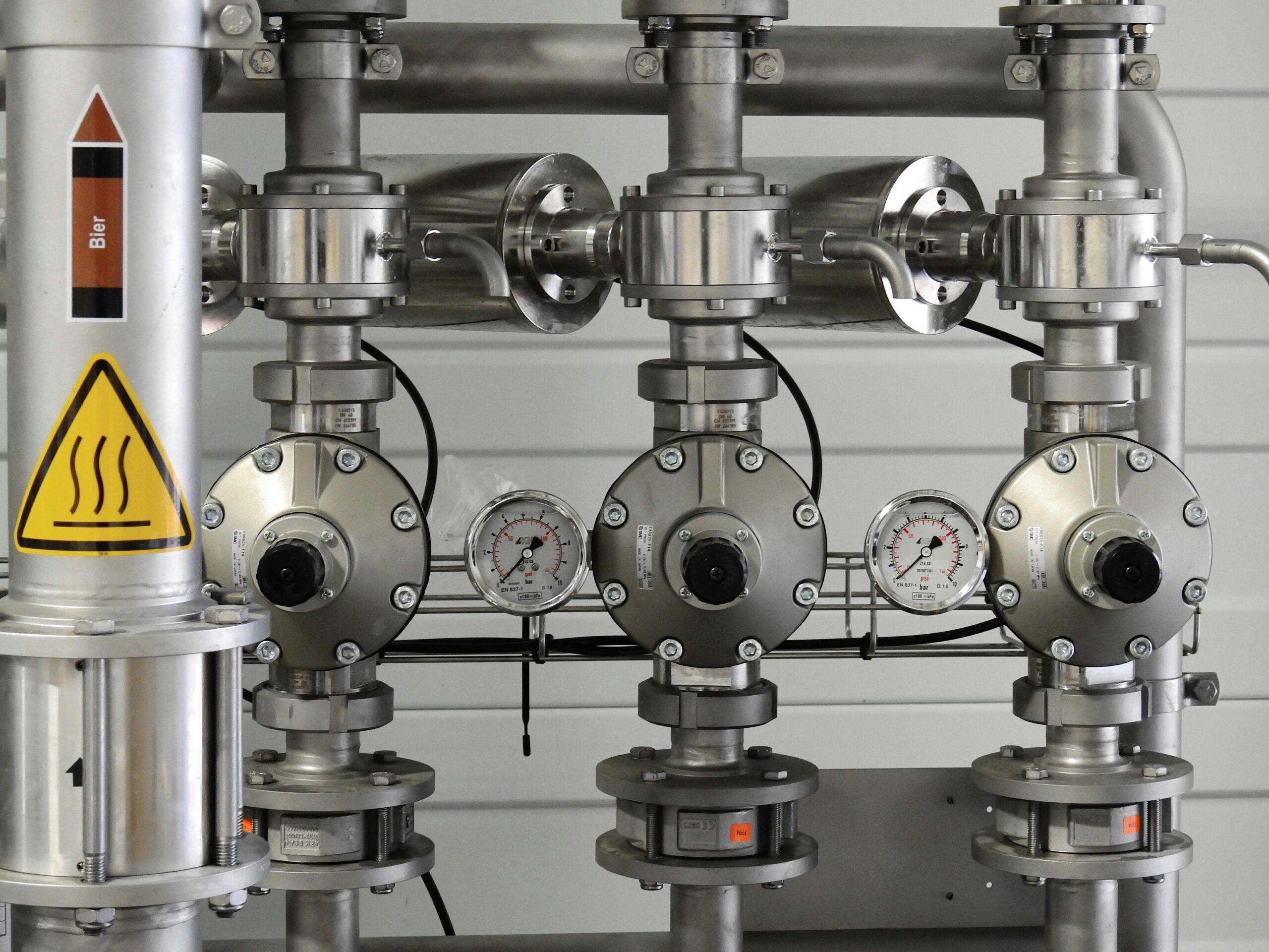 Qemi International Inc (2) A close-up view of an industrial piping system with stainless steel pipes, pressure gauges, and control valves. A warning sign with a heat symbol and the word "Bier" indicates the system is related to beer production or brewing. The gauges display pressure readings, highlighting the controlled process of fluid transfer.